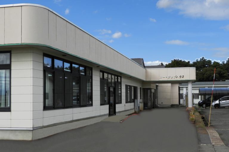 介護施設の建物外観。白い壁面と緑のアクセント、駐車場が見える。